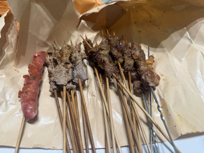 A photograph of various grilled meat skewers, including a sausage and pieces of beef and pork, resting on crumpled brown butcher paper.