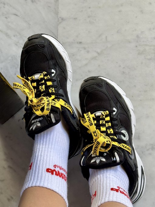 A pair of black and white sneakers with yellow laces printed with "BINANCE" text, paired with white socks, styled to visually reinforce the post's message of "moving forward, step by step, with Binance."
