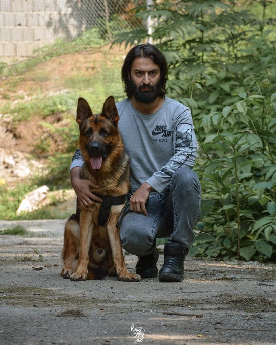 A man with long dark hair and a beard kneels beside his German Shepherd on a paved path surrounded by greenery, wearing a gray "NIKE AIR" long-sleeve shirt, jeans, and black boots. The image perfectly complements the post's sentiment about bonding more easily with other species than with humans, showcasing a moment of quiet companionship between the man and his loyal canine friend.