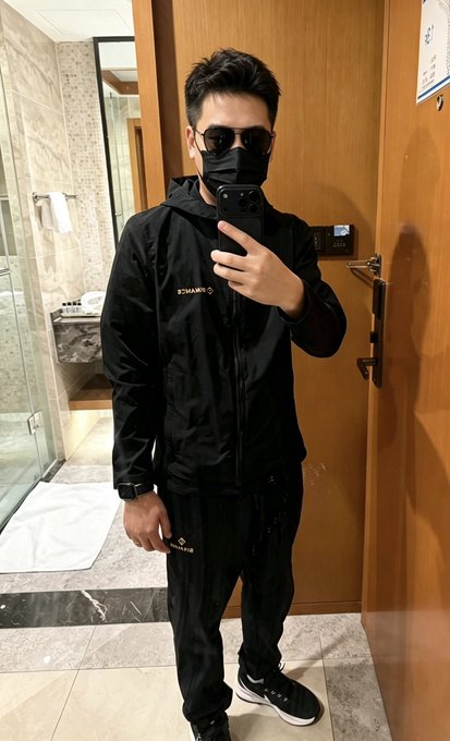 A man taking a mirror selfie wearing a black tracksuit, sunglasses, and a black face mask, standing in what appears to be a hotel bathroom.