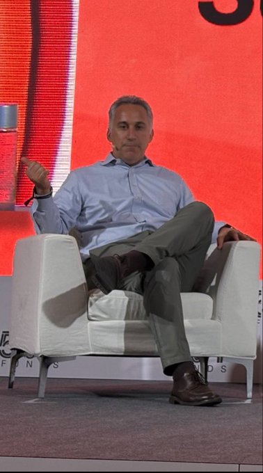 David Schamis, CEO of Hyperliquid Strategies ($PURR), sits casually on a white armchair on stage at DAS Asia, gesturing with one hand while wearing a light blue shirt and khaki pants during his panel discussion. The relaxed pose contrasts with the substantial details of his role managing an $888M HYPE treasury deal and a $2.5B equity facility expansion.