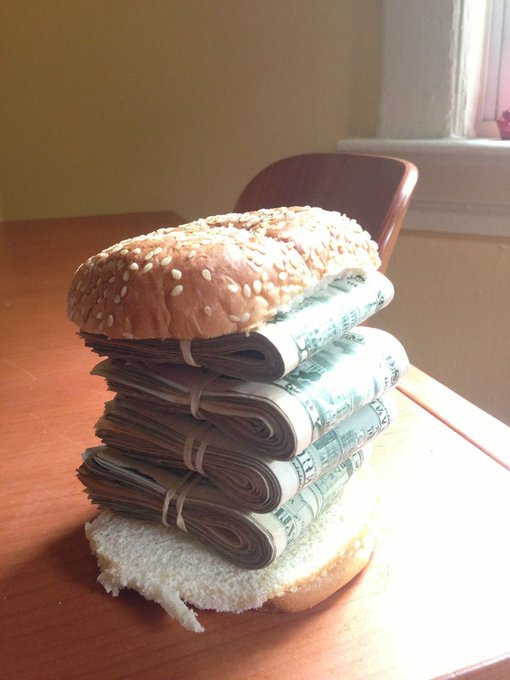 A sesame seed bun sandwich filled with stacks of rolled-up cash (hundred dollar bills), serving as a visual metaphor for a money burger.