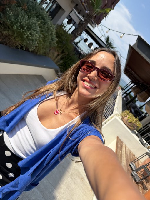 A woman takes a selfie outdoors, wearing red oval sunglasses, a white top, a blue shirt, and a pink heart necklace, with buildings and greenery in the background.