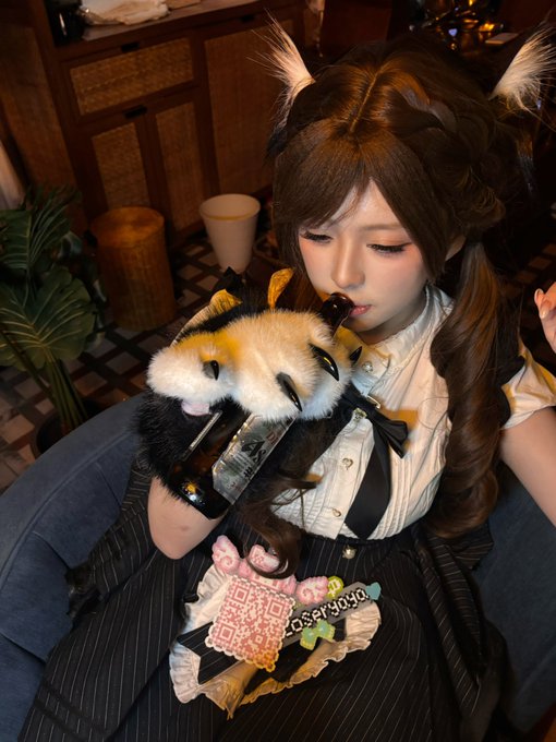 A cosplayer dressed in a black and white maid outfit with brown twin-tails holds a plush toy and a small bag, with a 'CroxyProxy' watermark visible on the image.