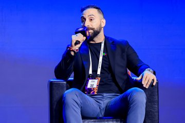 A bearded man in a dark blazer and jeans sits in a chair on a blue-lit stage, holding a microphone and wearing a green lanyard.