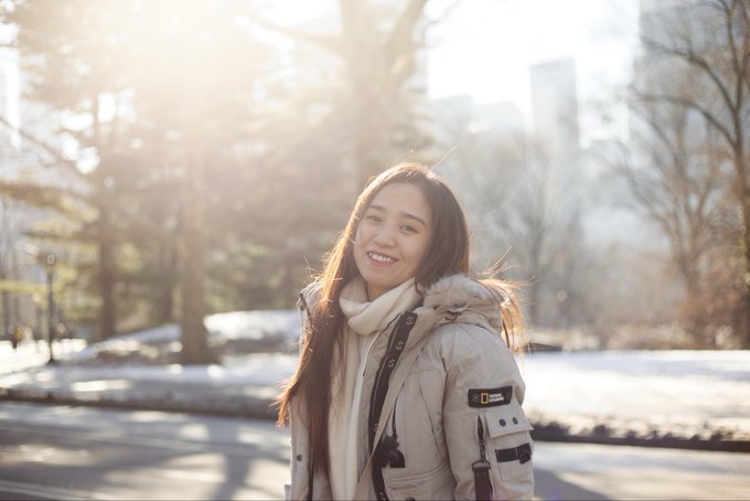 A smiling young woman wearing a beige puffer jacket and a white scarf stands outdoors in a park-like setting with snow on the ground and sunlight filtering through the trees.