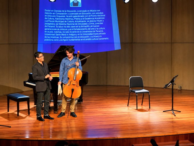 Two men standing on a stage next to a grand piano, one holding a cello, with a large screen behind them displaying Spanish text about 'Doctor en Ciencias de la Educación'.