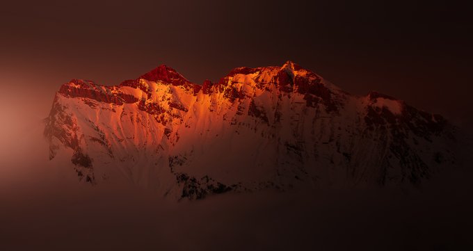 A snow-capped mountain peak glowing in dramatic red/orange light against a dark sky, likely captured during sunrise or sunset (alpenglow effect).