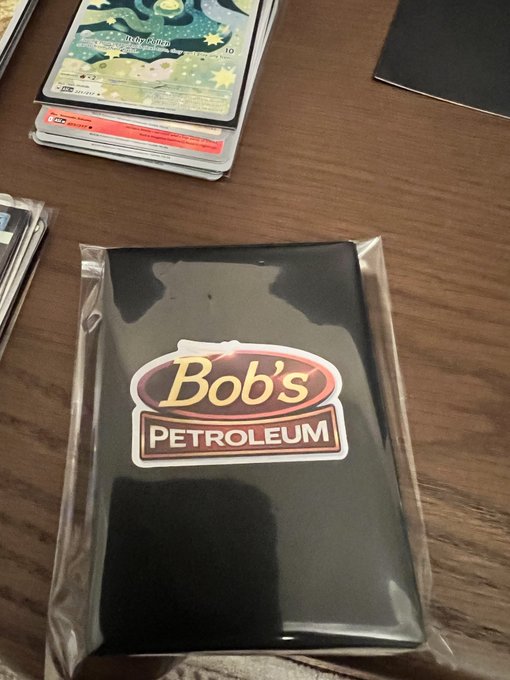 A sealed "Bob's Petroleum" card sleeve pack rests on a wooden table next to stacks of Pokémon trading cards, ready for its debut release.