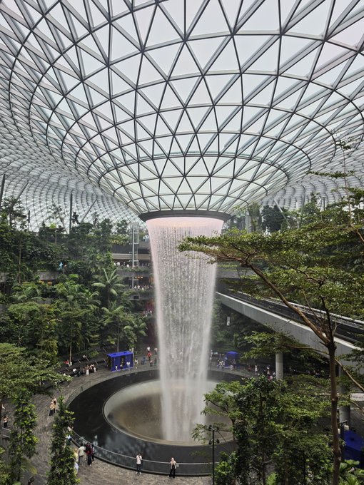 The interior of Jewel Changi Airport featuring the massive Rain Vortex waterfall surrounded by lush indoor gardens under a glass dome ceiling.