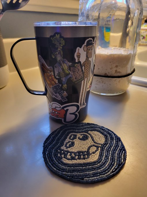 A black travel mug covered in stickers (including 'B' and various characters) sits next to a glass bottle with a brown beverage on a kitchen counter, accompanied by a beaded coaster featuring a skull design.