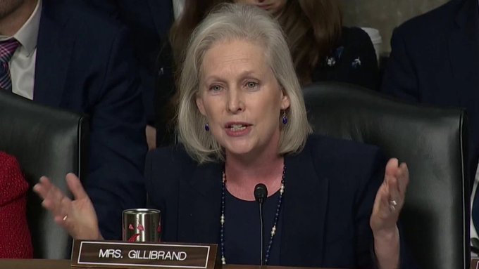 The image shows Senator Kirsten Gillibrand speaking with expressive hand gestures while seated at a hearing, with a nameplate reading "MRS. GILLIBRAND" visible in front of her. The photo relates to the post as it depicts the senator at the center of progressive backlash over her CLARITY Act ethics rules and alleged cryptocurrency conflicts of interest.