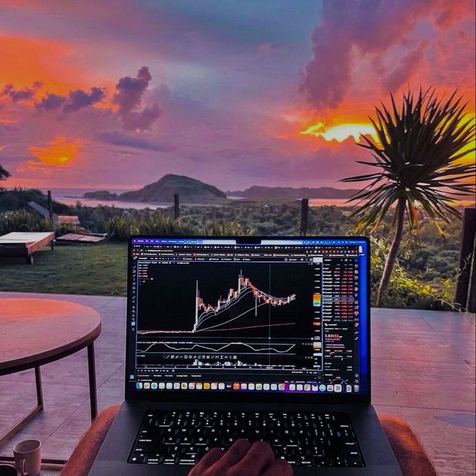 A laptop displaying a trading chart sits on a wooden table against a stunning sunset backdrop with vibrant orange and purple clouds over a coastal landscape.