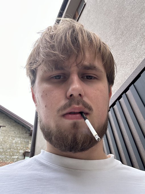 A close-up selfie of a young man with light brown hair and a beard, holding an unlit cigarette in his mouth.