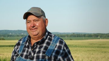 An older man wearing a plaid shirt, denim overalls, and a grey cap smiles while standing in a grassy field with trees in the background.