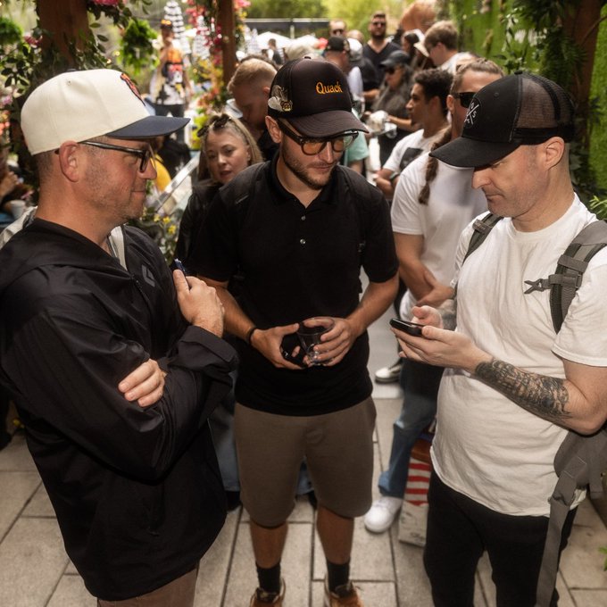 Three men wearing caps stand outdoors at what appears to be a social gathering, two of them looking down at their phones.