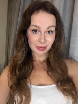 A selfie portrait of a woman with long brown wavy hair, wearing a white top, against a gray curtain background.