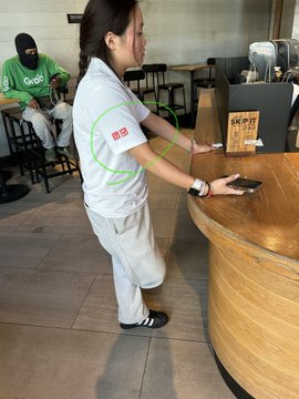 A woman wears a white Grab uniform featuring the company's name and green logo while working behind a wooden counter, illustrating the "status symbol" branding referenced in the caption. A man in the background also wears a green Grab shirt, reinforcing the company's visible presence.