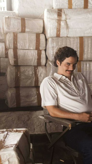 A man with curly dark hair and a mustache wearing a white polo shirt, seated in a chair and leaning against a wall stacked high with large wrapped packages or bales.