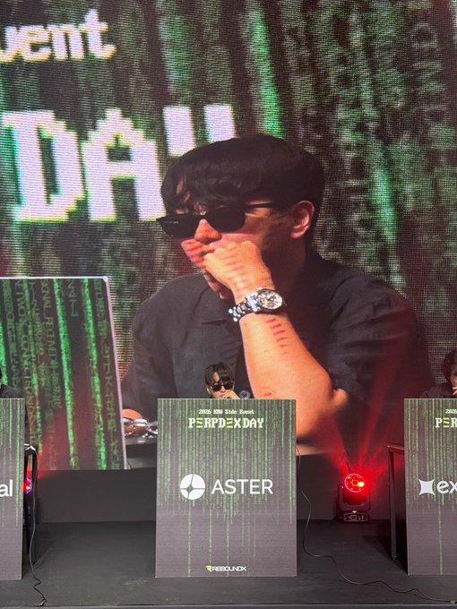 A man wearing sunglasses and a black shirt sits on stage at the 2025 ROK Slate Event "PERPDEX DAY," resting his chin on his hand in a thoughtful pose. He represents Aster DEX, as shown on the branded nameplate in front of him, connecting to the post about Aster DEX's presence at Korean Blockchain Week.