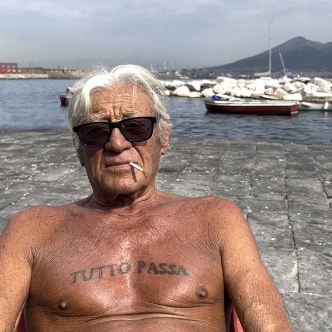 The image features an older man sitting outside near a body of water, wearing sunglasses and smoking a cigarette. The man has a tattoo on his chest that reads "Tutto passa", which translates to "I want to pass" in Italian. In the background, there are several boats docked at the pier, suggesting that this location is likely a popular spot for boating or water-related activities.