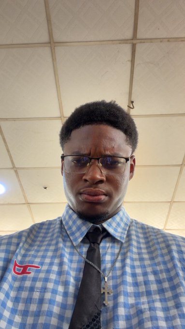 A young man in a blue checkered shirt, black tie, and glasses maintains a serious, unsmiling expression for the camera, fitting the "Day 1 of not smiling until $ANSEM hits $1" challenge. The drop-ceiling background and his resolute look emphasize his commitment to the pledge.
