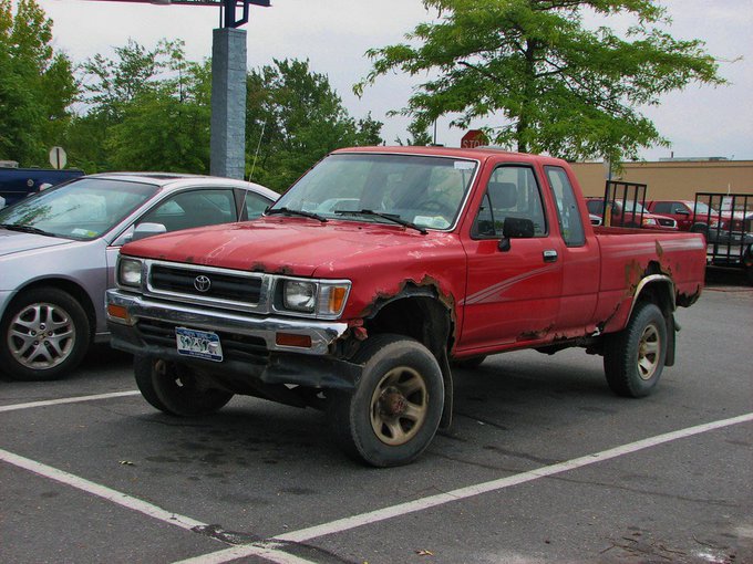 A beat-up red Toyota pickup truck with significant rust and body damage, particularly around the wheel wells, parked in a lot next to a silver sedan.
