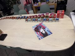 A light-colored table displaying an assortment of trading card game products, including booster packs, deck boxes, and a promotional poster in the foreground.