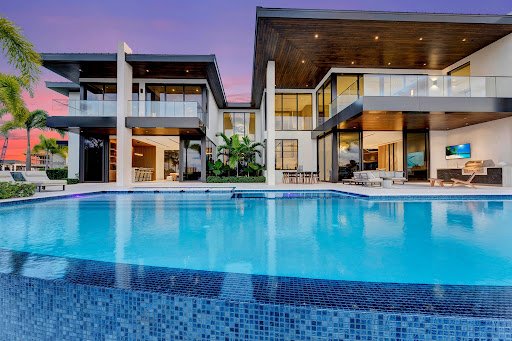A luxurious modern mansion at sunset with a large swimming pool in the foreground, featuring multiple stories, glass balconies, and elegant outdoor seating.