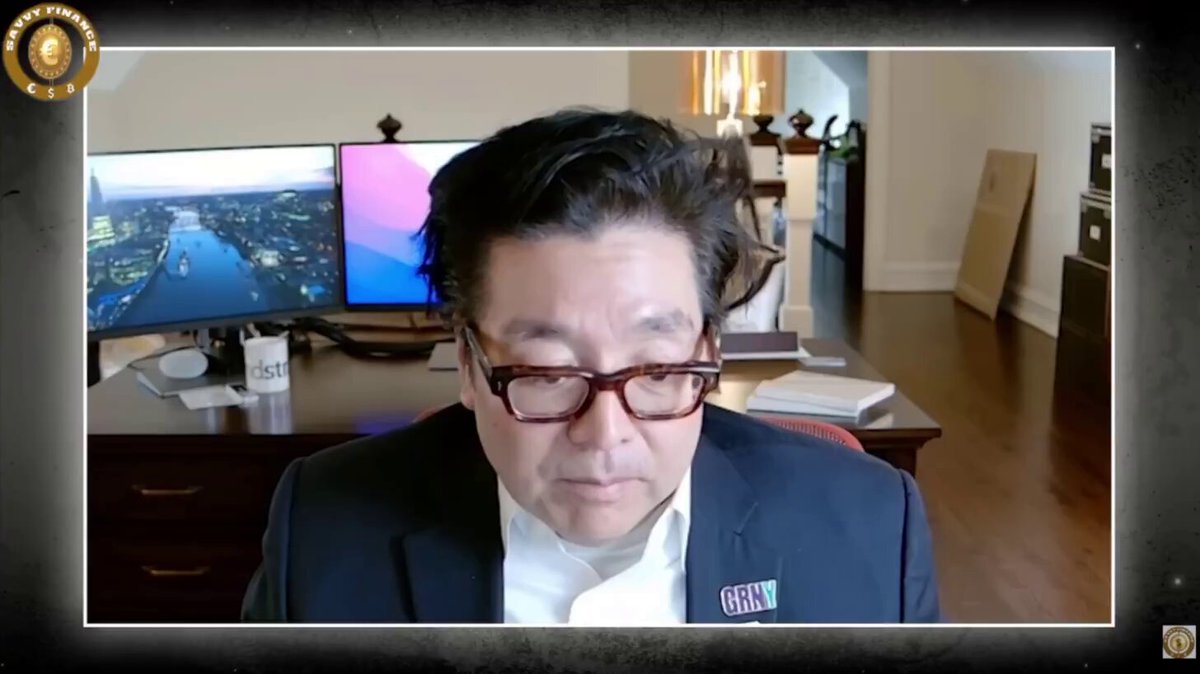 The image shows a man wearing glasses sitting at a desk with two computer monitors, one of which appears to be displaying a video call from another person. The man seems to be engaged in a conversation or presentation over the phone. The background features a wooden floor and a dresser, suggesting that he is likely in an office setting.