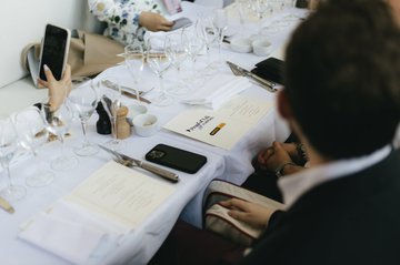 The image shows a group of people sitting around a long dining table at a restaurant. One person is using their cell phone while others are engaged in conversation or enjoying their meal. The table is set with wine glasses, plates, and utensils such as knives and forks. There is also a menu on the table for guests to choose from. The overall atmosphere appears to be social and relaxed, with people sharing food and drinks together.