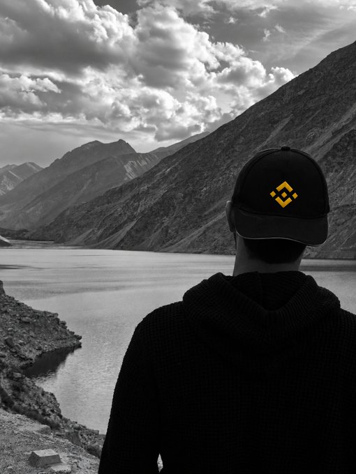A man in a black cap featuring a gold Binance logo gazes out over a black-and-white mountain lake, embodying the post's message of striving for excellence and rising above, with Binance standing as a symbol of elevated standards in the crypto world.