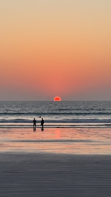 Two silhouetted figures stand on a beach at sunset as the orange sun dips toward the ocean horizon.