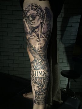 A detailed black-and-gray tattoo sleeve on a person's arm featuring a figure with a laurel wreath, sunglasses, and Roman-style elements, with a banner reading 'HIMS' across the forearm.