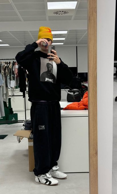 A mirror selfie shows a person wearing a yellow beanie, a black graphic sweatshirt, black track pants, and sneakers, standing in what appears to be a clothing store.