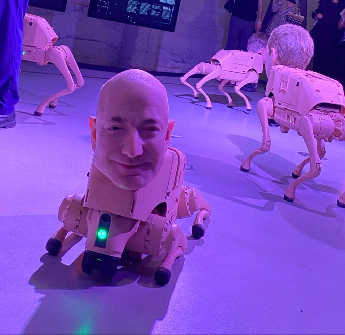 A bald, smiling man's face appears digitally edited onto a small robotic quadruped body, displayed in a purple-lit exhibition space with several similar robots and attendees visible in the background.