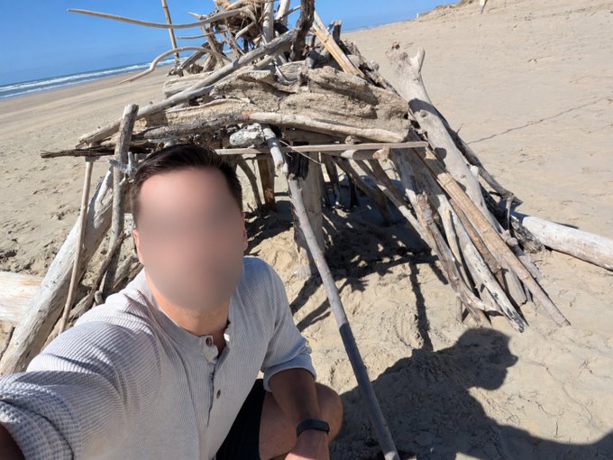 A selfie of a person with a blurred face sitting on a sandy beach in front of a large structure made of driftwood.