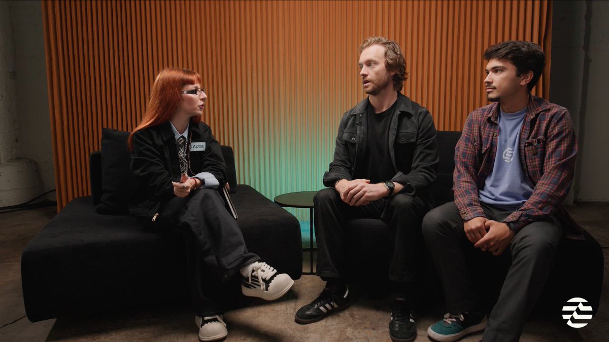 The image shows three people seated in what appears to be an interview or discussion setting, with a red-haired woman wearing an "Aptos" name tag conversing with two men—one in a black jacket and another in a plaid shirt—against an orange curtain backdrop with green lighting accents. The scene visually corresponds to the post's description of a conversation between PACT Co-Founders Zander Rafael and Eric Cuellar with the Aptos Foundation's representative, discussing Tether's $7M investment in PACT Labs to integrate USAT into payroll and payment systems.