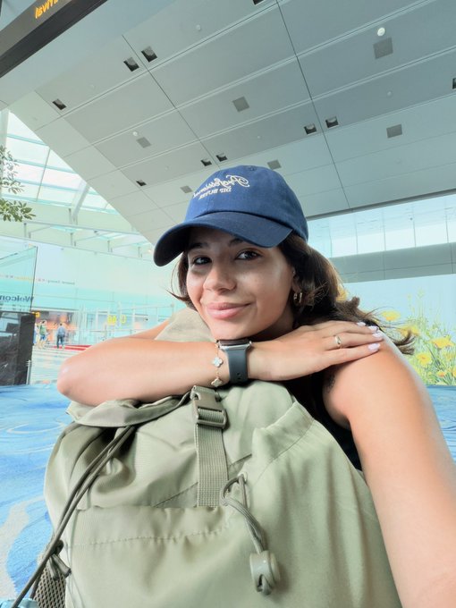 A young woman wearing a blue baseball cap and carrying a green backpack, taking a selfie inside what appears to be an airport terminal.