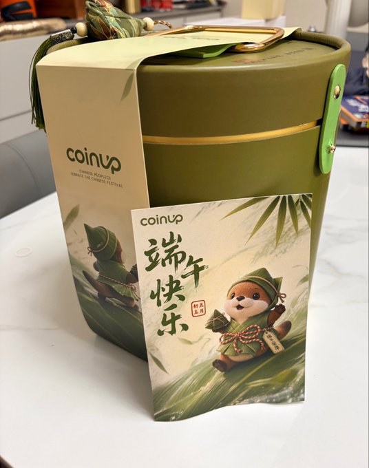 The image features a green container with Chinese characters, possibly a bucket or tin can, sitting on top of a white table. The container has a label that says "CoinUp" written in English and Chinese. There are also some Chinese characters visible on the side of the container. A small note is placed next to the container, which reads: "礼盒啦", meaning "I want to send you".