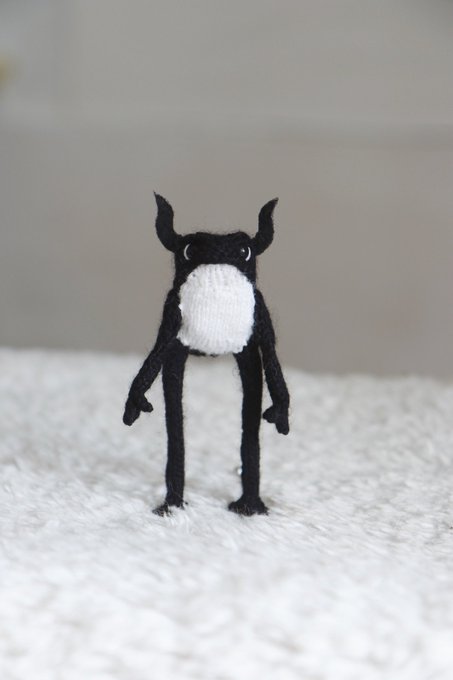 A small handcrafted black and white felted monster figurine with horns, round white belly, and stick-like limbs standing on a white textured surface.