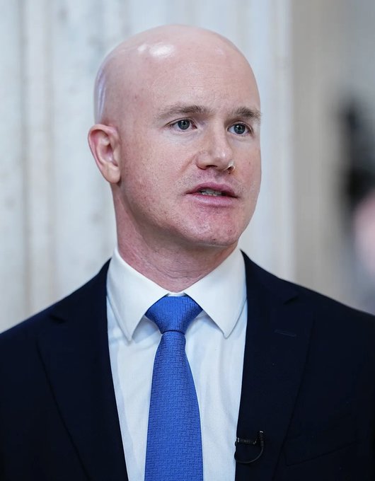 A bald man in a dark suit, white shirt, and blue tie speaks to reporters, with a small lapel microphone clipped to his jacket. The image appears to accompany a post about Brian Armstrong, CEO of Coinbase (which operates the Base blockchain network mentioned in the text regarding the $LAPTOP meme token's launch).