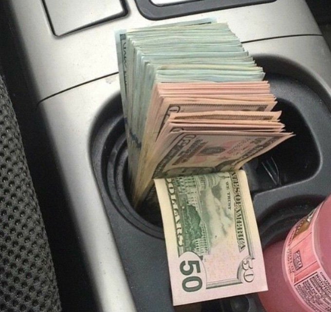 A thick stack of U.S. $50 bills, fanned out to reveal multiple "50" denominations, is crammed into a car's cup holder beside a bottle of what appears to be cleaning product. The image visually represents the post's aspiration of "making money at a young age," showcasing a tangible display of cash wealth as a symbol of financial success.