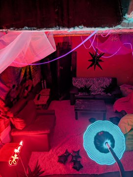 The image shows a living room with red walls, pink curtains, and various pieces of furniture such as couches and tables. There are also neon lights hanging from the ceiling that create an interesting atmosphere. The photo has been edited to include a caption about a party or event happening at Berlin Blockspäti next week.