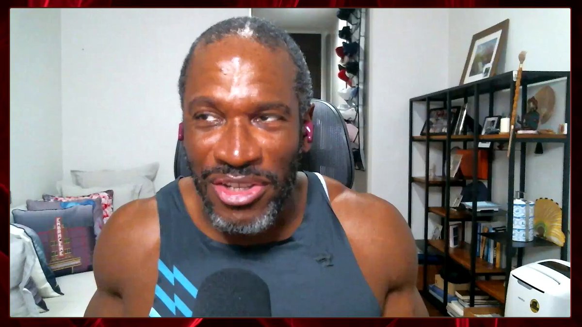 The image shows a man wearing a black tank top with blue stripes, sitting in front of a computer screen during an interview. The man appears to be talking about his thoughts on the current state of cryptocurrency markets or AI technology. The background reveals that he is in a room filled with books and other items, suggesting that this could be a home office or a workspace.