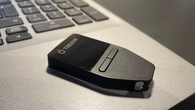 A black Trezor hardware wallet sits on a laptop next to a keyboard, with the Trezor logo visible on its surface. The image illustrates the device at the center of a recent security concern, where Trezor warned approximately 13,689 customers about a data breach at a shipping provider that may have exposed their personal information.