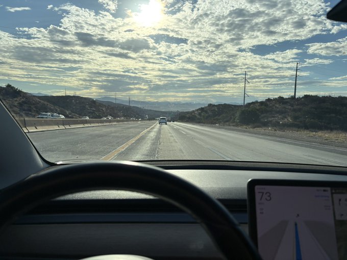 A view from inside a moving vehicle, looking through the windshield at a multi-lane highway under a bright, cloudy sky, with the dashboard display showing a speed of 73 mph.