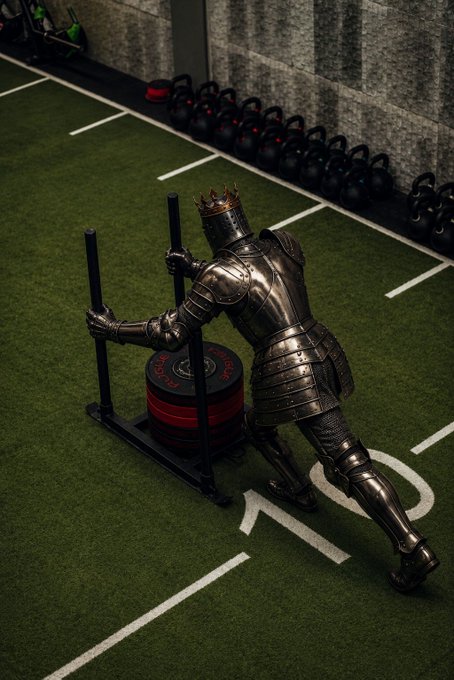 A knight in full silver armor wearing a crown pushing a weighted sled loaded with bumper plates across artificial turf in a gym, with kettlebells lined up against the wall.
