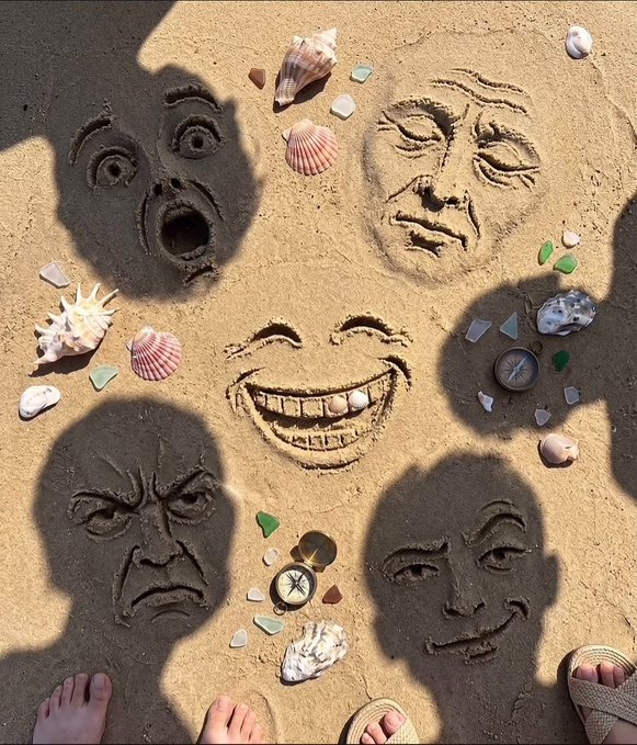 An overhead photo of faces drawn in sand surrounded by seashells, sea glass, and compasses, with the shadows of people standing around the artwork.