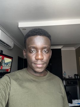 A young man in an olive green t-shirt takes a selfie indoors, with a television visible in the background.